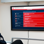 **

A brightly lit office scene. A diverse group of employees are attentively watching a security awareness training presentation on a large screen. The presentation slide features examples of phishing emails with red flags highlighted. Several employees are nodding in understanding. The overall tone is positive and proactive. In the background, a poster with the tagline "Güvenlik Seninle Başlar" ("Security Starts With You") is visible.

**